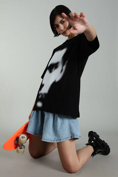 Model in oversized black t-shirt with abstract face design, kneeling and holding a skateboard, showcasing urban fashion.