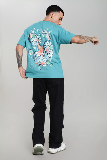 A man wearing a teal unisex oversized fish t-shirt with a colorful fish design on the back, standing against a plain background.