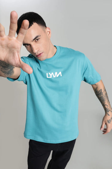 A man wearing a teal fish unisex oversized t-shirt with a logo, making a stop gesture against a gray background