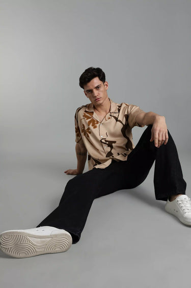 Man sitting on a gray background wearing a beige boho shirt and black pants.