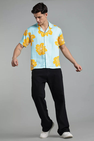 Man wearing a light blue shirt with yellow floral patterns and black pants on a gray background