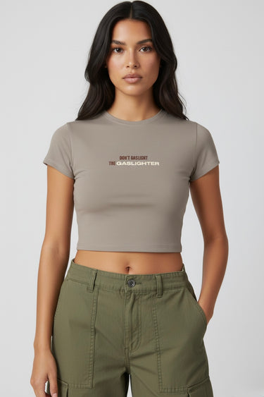 Woman wearing a dark beige crop top that says "Don't Gaslight the Gaslighter" and green pants on a white background