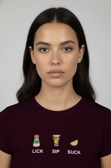 Woman wearing a dark maroon graphic crop top with graphic designs and text 'LICK', 'SIP', 'SUCK' on a plain background