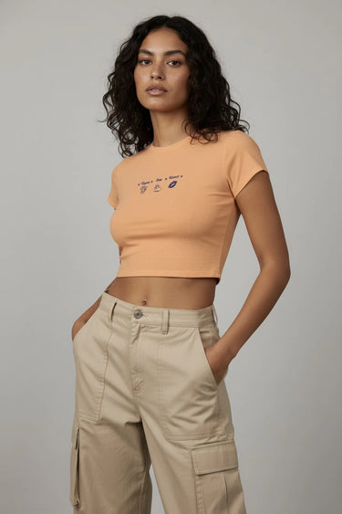 Woman wearing an orange graphic crop top with text and beige cargo pants against a gray background