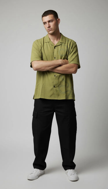 Man wearing a relaxed fit green embroidered shirt and black pants standing with arms crossed on a gray background