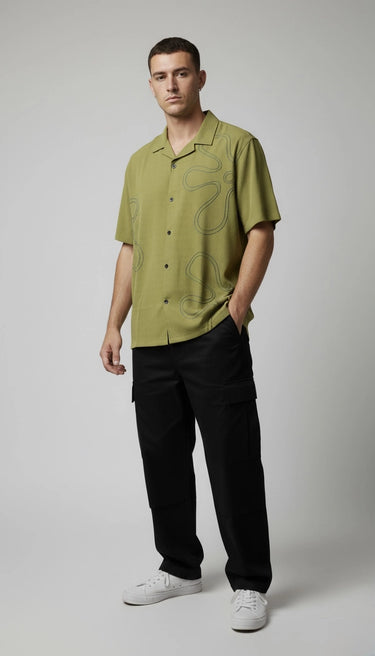 Man wearing a green relaxed fit embriodery shirt and black pants on a gray background