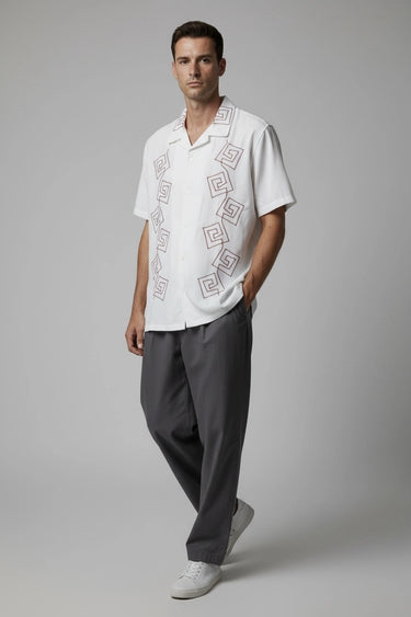 Man wearing a white embroidered relaxed fit shirt and gray pants on a plain background