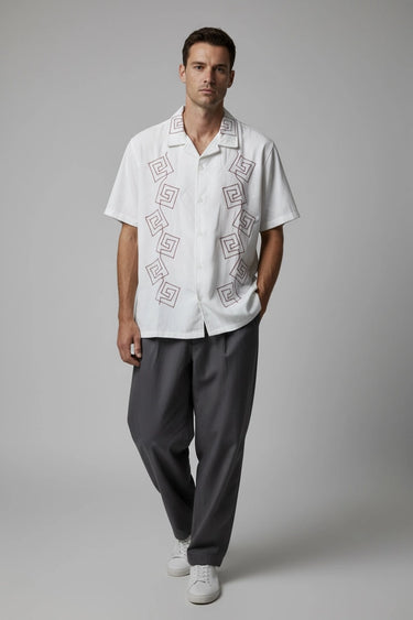 A man wearing a white embroidered relaxed fit shirt and gray pants on a plain background