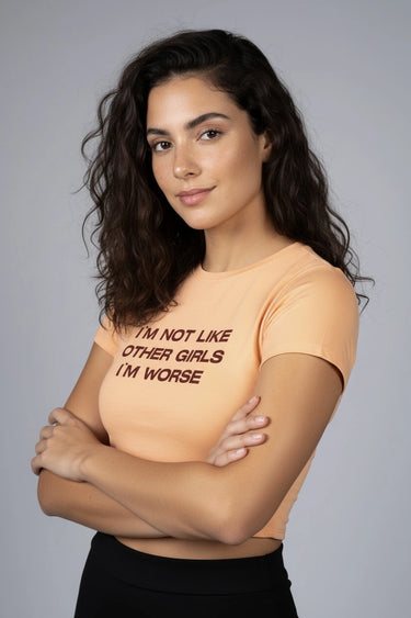 Woman wearing an orange graphic crop top that says "I'm not like other girls I'm worse", standing against a gray background