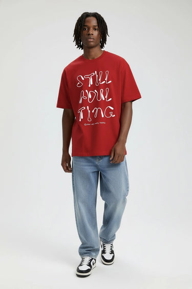 Red oversized graphic t-shirt with "Still Adulting" text paired with light blue jeans and black and white sneakers.