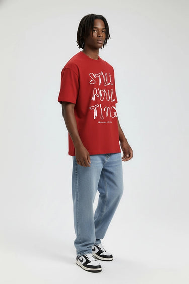 Red oversized graphic t-shirt with "Still Adulting" white text, worn with light blue jeans and black-and-white sneakers.
