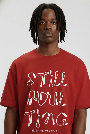Red oversized graphic t-shirt with bold white "STILL ADULTING" text and loose fit on model.