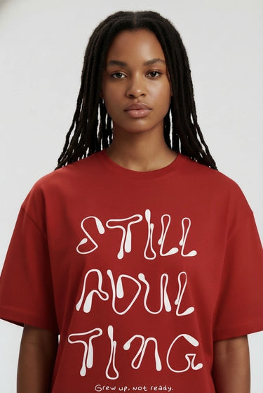 Red oversized graphic t-shirt with white "STILL ADULTING" text worn by woman.