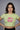 Woman wearing a green GRAPHIC crop top with pink text that says is she me against a gray background
