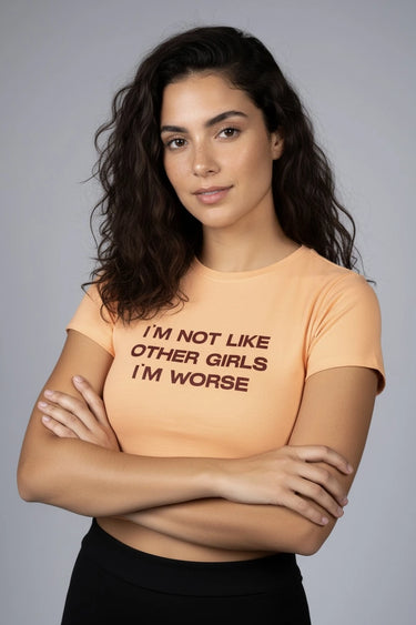 Woman wearing an orange graphic crop top with text, "I am not like other girls  I'm Worse" standing against a gray background 