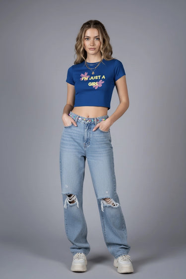 Woman wearing a blue graphic crop top with text that says "I'm Just a Girl" and light blue jeans on a gray background